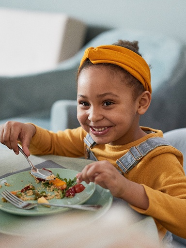 Little girl in yellow shirt eating food from plate with fork and spoon