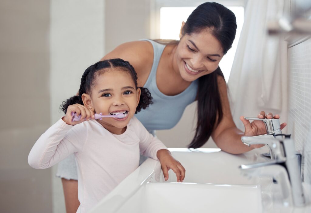Woman with dark hair helping little girl brush her teeth at bathroom sink